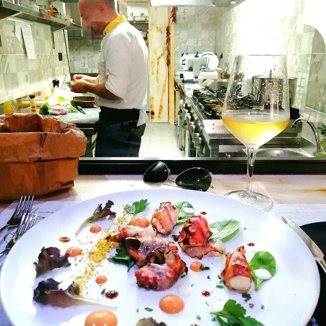 Calice di vino in primo piano, cucina aperta di Serrago sullo sfondo